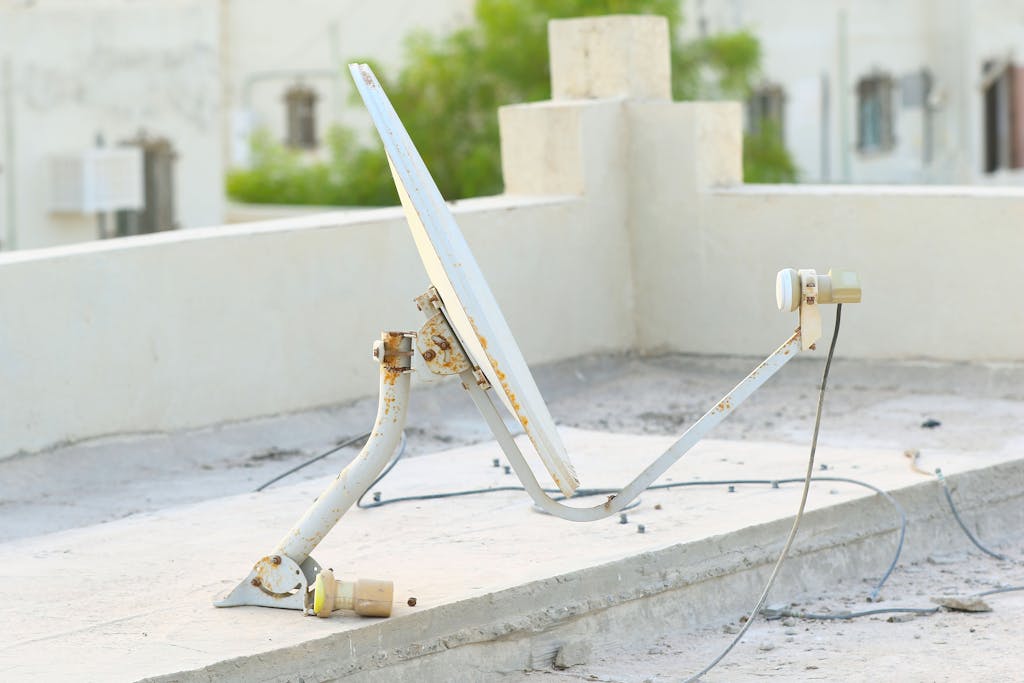 Old rusty satellite dish on a concrete rooftop, surrounded by wires and urban scenery.